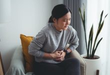 Asian woman feeling stomachache and touching her belly sitting on armchair in living room at home.