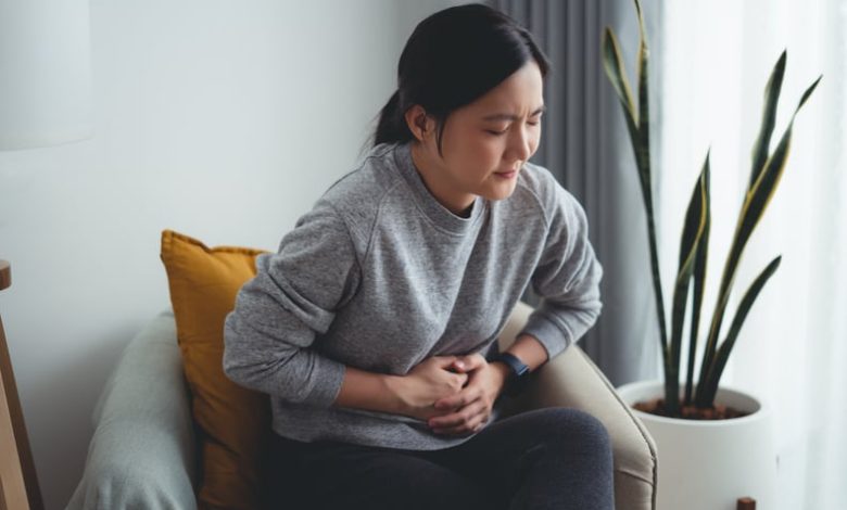 Asian woman feeling stomachache and touching her belly sitting on armchair in living room at home.